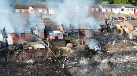 An aerial shot of a row of homes with their roofs destroyed by fire. Smoke streams into the air. Land in front of the houses is scorched, with firefighters holding hoses spraying water towards the scene.