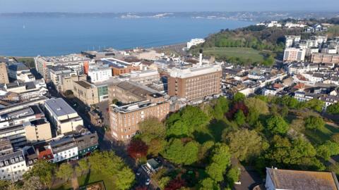 An aerial view of a hospital site, with tall brick buildings surrounded by trees on one side and the sea on the other.