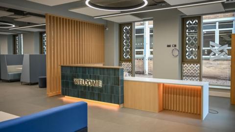 A reception desk in a newly-built building. Part of it is covered in green tiles with "welcome" in gold letters. A seating area can be seen on the left and there are two windows behind the reception desk.