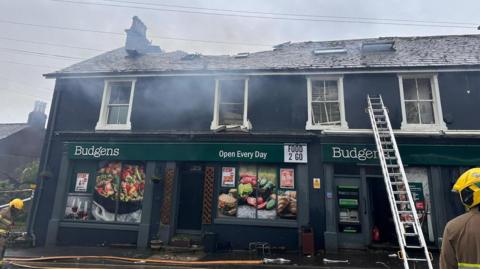 A Budgens store with fire damage to the roof, a fireman's ladder to the left of the scene, and smoke haze in the air.