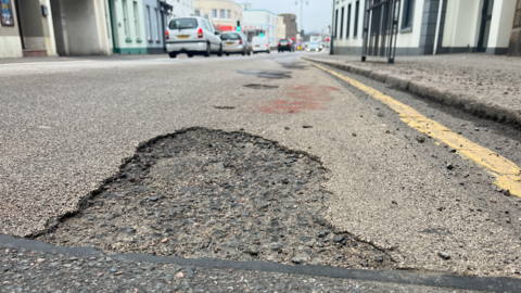 A pot hole on a main road with traffic on the left in the background.