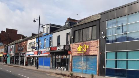 Holmeside has two-storey properties, with takeaways, a barber shop and a repair shop on their ground floors. Dr Q's Food Factory is also in the shot, with its blue shutter down. 