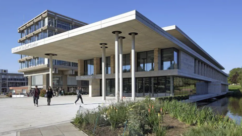 A picture of two of the modern buildings at the University of Essex. One of the concrete buildings is next to a lake, which is on the right of the picture. Three students are walking across a large, paved forecourt. Other buildings on the campus can be seen in the distance. It is a sunny day.