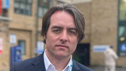Thom Lafferty has long brown hair and is wearing a dark suit jacket over a white shirt. The beige brick hospital building is behind him. He looks serious in his expression.
