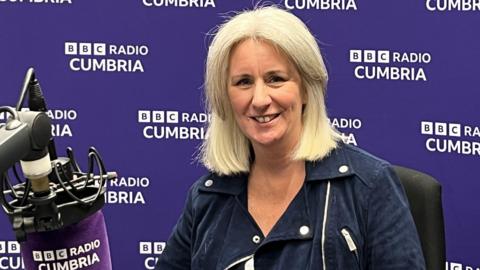 Heather Wainwright sitting in a BBC Radio Cumbria studio, with a branded purple microphone in front of her and a branded purple wall behind her. She has blonde/white hair in a bob and is wearing a blue jacket. She is smiling at the camera.