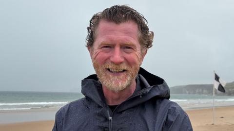 Andrew Todd stands on Whiterocks beach on Portrush. He has wavy brown hair and a ginger/grey beard. He is wearing a dark blue raincoat. It is an overcast day.