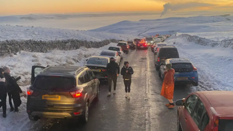 Narrow road surrounded by snowy landscape with roads parked either side of the road and some cars driving down the middle