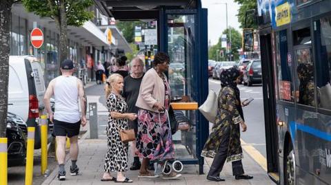 People at a bus stop on Kings Heath High Street in Birmingham queue to get on a bus. Three women are holding bags, a trolley and a phone and two men are wearing shorts. A bus is pulled up close to the pavement. 