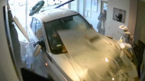 Silver car crashed into an office, with a shocked woman in the background.