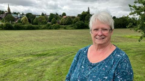 Elizabeth Scott with short white hair, glasses and a blue top with a nature print on it, stands in front of a vast field bordered with trees