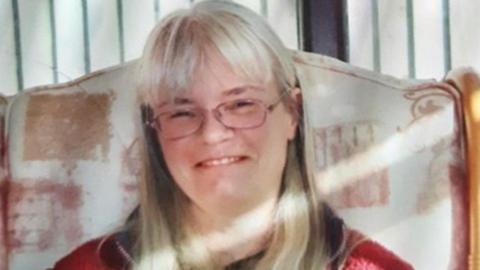 A lady with shoulder length grey hair and glasses is sat on a large chair with gold sides and smiling at the camera. She is wearing a red jumper.