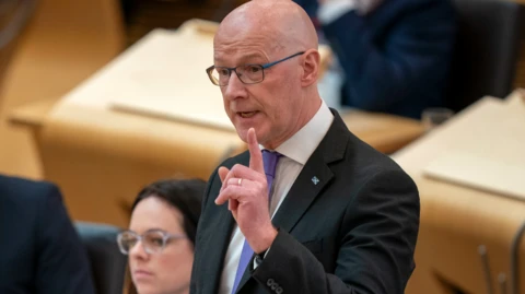 John Swinney, a bald man with glasses and wearing a dark suit, purple tie and white shirt, points a finger in the air as he addresses MSPs