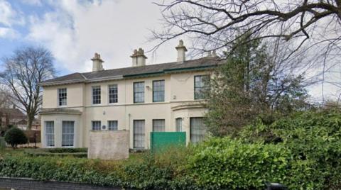 A light-coloured two-storey building has a tree to the left of it on the photo. A hedge is above a small brick wall in the foreground.