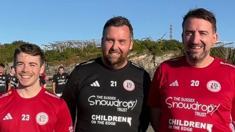 Joe Hepworth, Angus Stewart and Mark Oliver wearing their football jersies. They are standing at the Henningsvaer Stadium in Norway. Behind them are their friends.