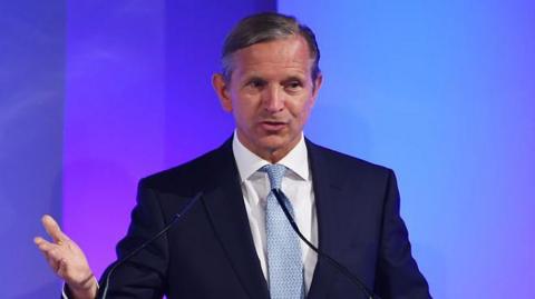 A man wearing a navy blue suit, white suit and pale blue tie gestures with his right hand as he speaks into two microphones - he has short, dark grey hair and is in front of a purple and blue backdrop 