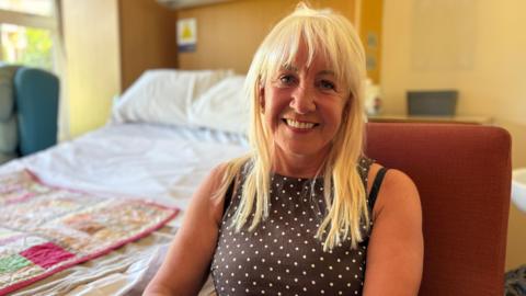 Alison has light hair and is wearing a black and white polkadot top. She smiles at the camera with a 'cuddlebed' behind her. 