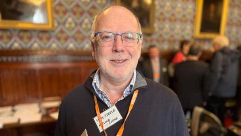 Mark Steele in a dark jumper and glasses, wearing an orange lanyard and name tag, standing inside the Houses of Parliament in Westminster