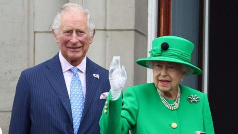 Prince Charles in a blue pinstripe suit standing beside the waving queen in a green hat and dress