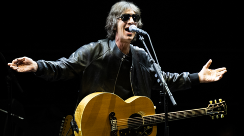 Richard is on stage with his arms open wide and palms facing up as he sings a song. He has a guitar over him and is wearing a black leather jacket and shades. He has floppy, brown hair.