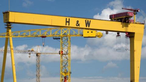 A large yellow crane stands against a blue sky with a few clouds. There are large black letters on them - H & W.