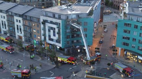An aerial photo showing eight fire engines stopped in a town square, with a block of flats on the corner blackened by fire. There's smoke still rising from the building, with a ladder rising from one of the fire engines.