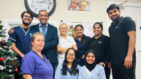 Staff at the care home posing for a group shot. Six of them are standing up and three are on their knees in front of them. They are all smiling. There is a Christmas tree to the left of the picture.