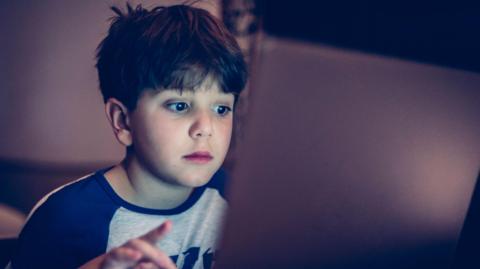 A young boy is looking at a laptop with his finger on the mouse pad. He is wearing a blue and white long-sleeved shirt.