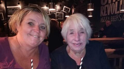 Two women smile in a bar while holding drinks. The younger woman on the left wears a purple top, and the older woman on the right wears a navy jacket and top.