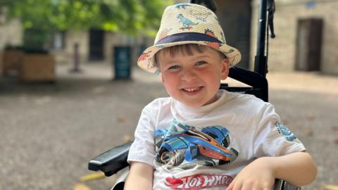 A smiling little boy wearing a dinosaur sun hat and a white t-shirt sits in a black wheelchair