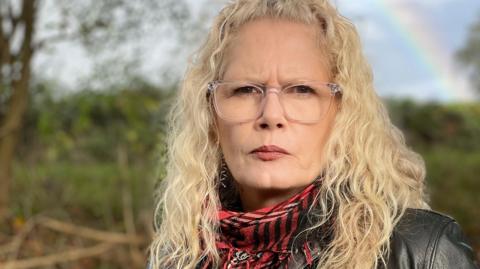 Head and shoulders photo of a woman with long curly blonde hair and clear framed glasses. She is wearing a black leather jacket and tartan scarf. The field in the background and part of a rainbow in the sky is blurred behind her.