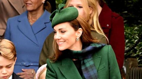 Close up of Kate Middleton's smiling face. She is wearing a green hat and coat and is stood close to her oldest son George.