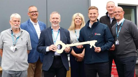 Dr Francis Old, Chris Couch, Steve Williamson, Lynda Sherfield-Wong, Karl Martin, Jeff Houghton and Lewis Saundry are standing in a line smiling outside the building. Mr Williamson is holding a giant key.