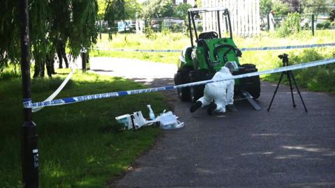 A person in white overalls is on the ground at the front of a small, green, single-seater, four-wheeled vehicle which has two forks at the front. It has a roof over the driver's seat. It is cordoned-off on a path, and has blue and white police tape around trees.