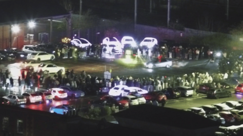 Grainy footage showing dozens of cars in a car park. A large crowd of people are gathered in a circle, which has two cars in the middle. One of the cars appears to have smoke coming out of the back of it.