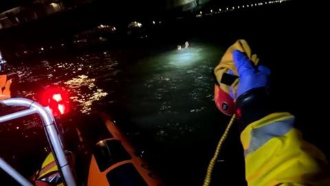 Edge of orange lifeboat with yellow sleeve and blue-gloved hand holding a yellow rope a light shines on the dark sea with the head of a person in the water illuminated