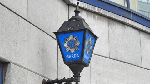 A close up of a lantern outside a garda station. The gardaí logo is on the lantern, which is attached to a grey brick wall. 