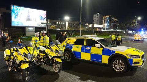 Police bikes and a car blocking the road close to the bus station with flats in the background. It is nighttime. Officers stand around wearing hi vis.