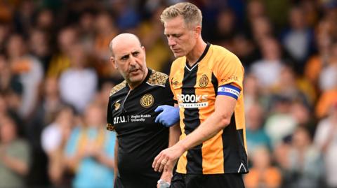 Michael Morrison walks off the pitch with a member of Cambridge's medical staff
