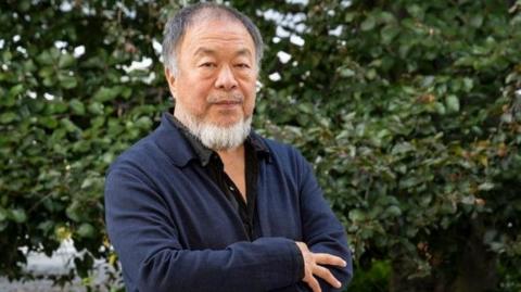 Ai Weiwei, who is wearing a black shirt and a navy overshirt, stands in front of a hedge with his arms crossed. He has a grey beard. 