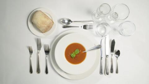 A bowl of soup on a white table cloth surrounded by a full set of cutlery