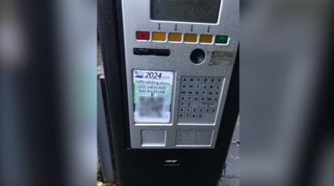 In an attempt to scam drivers, a ticket machine in a Luton car park has a sticker stuck over it's instructions featuring a bogus QR code. The box has a black surround and a silver face with multi-coloured squares along with letters of the alphabet and a white piece of paper with a black and white QR code on it.