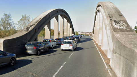 The Heugh Street Bridge has two lanes, with spaces for parked cars on the edge of the left lane, where about 10 vehicles are parked. A concrete arch with pillars on each side of the bridge separates the road from the pavement.