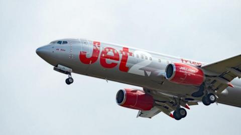 A Jet2 aeroplane flying against a pale grey sky. It is painted white and has Jet written in red and 2 written in white in large letters on its body