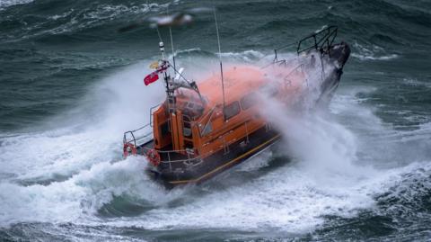 A Shannon lifeboat is in rough and heavy swell. A wave is breaking over it and its bow is lifted out of the water. It has a navy hull and orange wheelhouse.