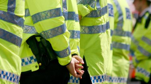 The backs of six police officers standing in a line. They are all wearing their hi-vis uniforms and one is standing with his hands linked behind his back.