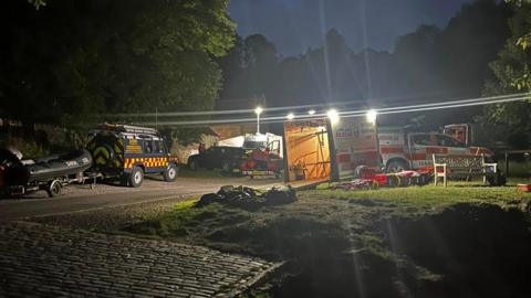 Lifeboats and emergency service vehicles are parked on the edge of a river bank. Floodlights are being used to assist the search.