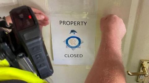 A police officer taping an A4-size "Property Closed" paper sign on a door. The officer's arms and radio are visible along with a section of a yellow hi-vis top. The door is painted white and has a gold coloured handle.