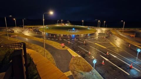Newby West roundabout at night. There are roadwork cones all along the road, which has been newly resurfaced. The image shows the completed roundabout, but with the road closure still in place.
