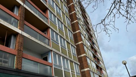 Windows and balconies of a rundown 1960s tower block in Gloucester, which is rising against the grey sky. Bare branches can be seen hanging down from a tree. The tower block is mostly brick with green cladding on the windows and glass and red cladding on the balconies.