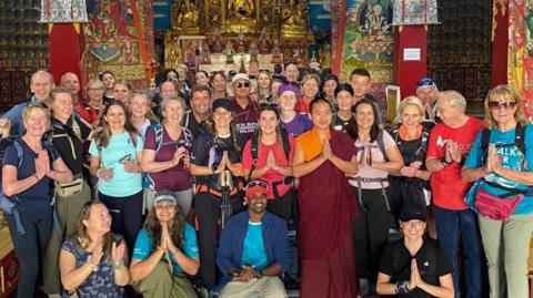 A large group of people in hiking gear stood infront of a buddhist temple with their hands together in prayer and smiling at the camera